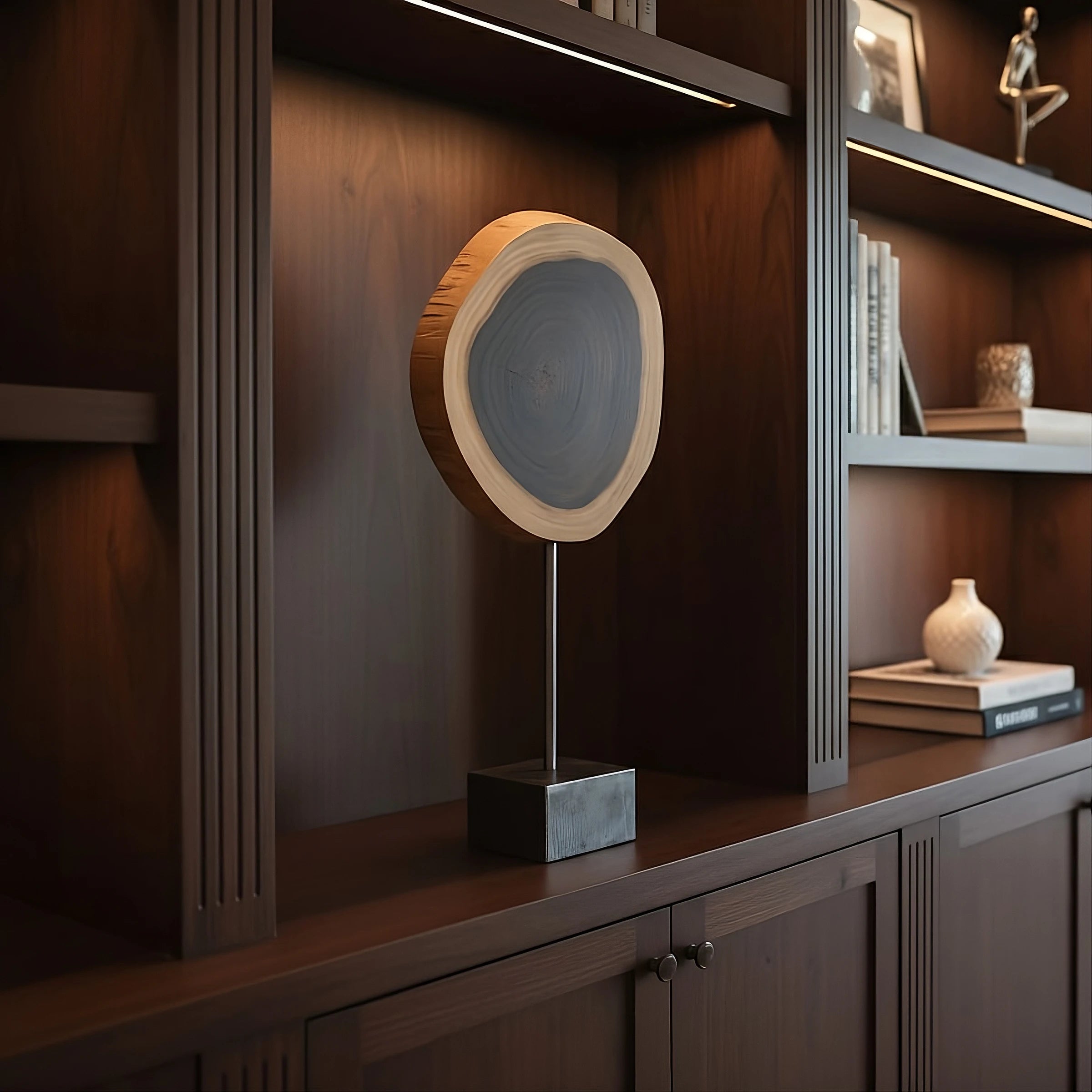 Cocuswood is a modern tabletop sculpture featuring a polished, natural wood slice mounted on a textured stainless steel base. The wood element is elevated on a slender, cylindrical metal rod that attaches to the base.