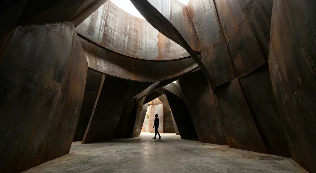 Large-scale sculptural installation made of weathered metal panels forming an enclosed, immersive architectural space. Silhouette of a person for scale.