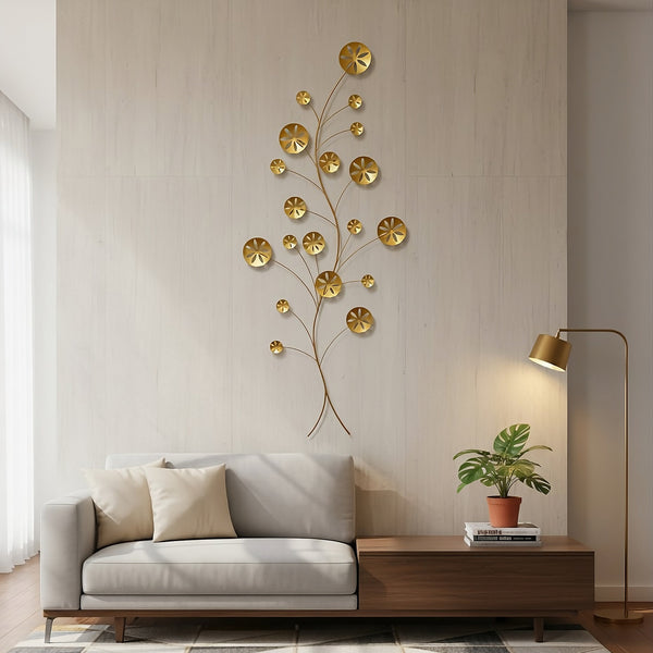 The gold "Gold Coast" metal wall sculpture, approximately 40 inches high, installed vertically above a light gray modern sofa in a living room with pale, textured walls. A low, dark wood console table sits next to the sofa, illuminated by a tall, gold-toned floor lamp.