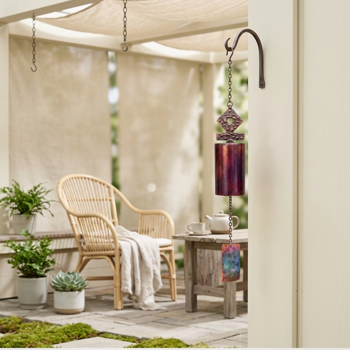 A hanging wind chime with a purple and red gradient cylindrical body and a blue patterned windsail, displayed on a beige patio next to a wicker chair and small table.