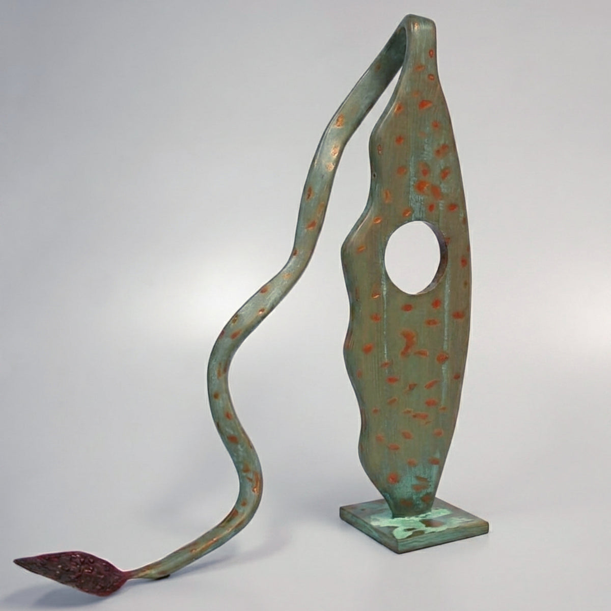 Modern abstract metal sculpture, featuring a verdigris green body with scattered orange speckles and a large circular cutout, viewed from a three-quarter angle on a neutral grey studio background. The slender main body tapers to a curved, sinuous arm ending in a small, textured, deep red leaf-like point resting on the ground, all anchored by a small, patinated square base.