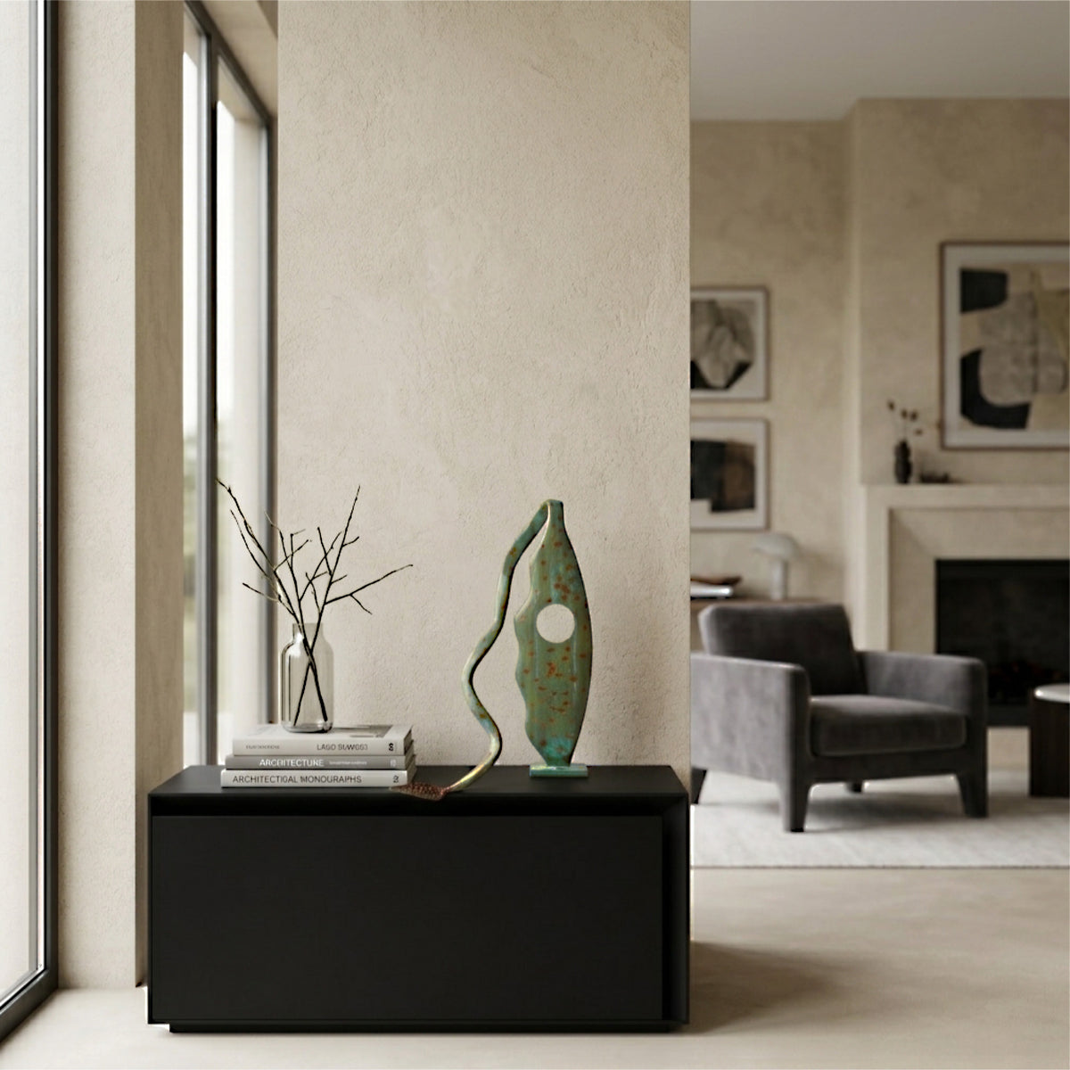 Modern abstract metal sculpture with a mottled green verdigris and terracotta patina, displayed on a low matte black console beside minimalist branches in a clear glass vase, set within a sunlit, contemporary interior with warm plaster walls and tall windows.